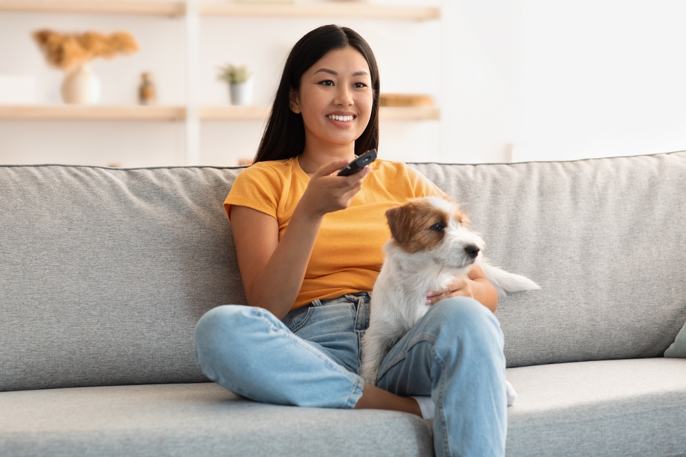 A woman sits with her Jack Russell Terrier puppy on the couch while she holds a TV remote.