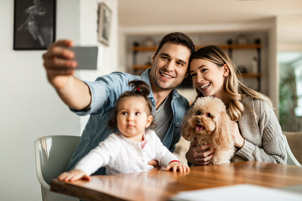 A beautiful, smiling Spanish family takes a selfie with their Miniature Poodle in the living room.