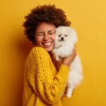 A woman wearing an orange sweater holds a white Pomeranian dog and smiles at how cute her furry friend is!